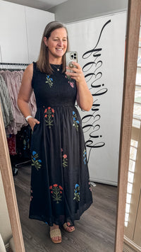 A woman takes a mirror selfie in a boutique showing a sleeveless black maxi dress with colorful floral embroidery. The boutique has a white wall with black script text and clothing racks are visible in the background.