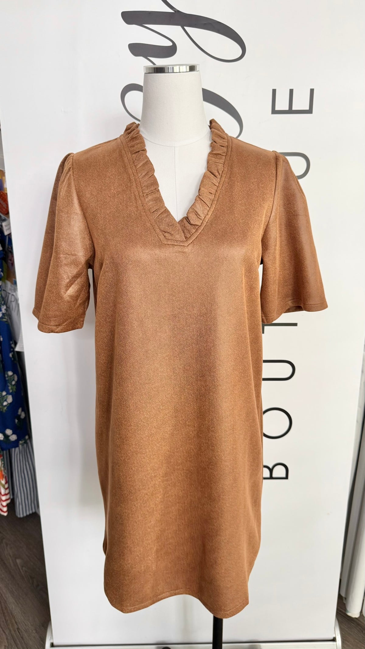 A tan short-sleeve V-neck dress with ruffled trim, displayed on a mannequin in front of a white boutique sign.