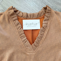 A close-up of a brown ruffled V-neck top with a white brand label reading 'bizzy lizzy BOUTIQUE' on a gray wood background.