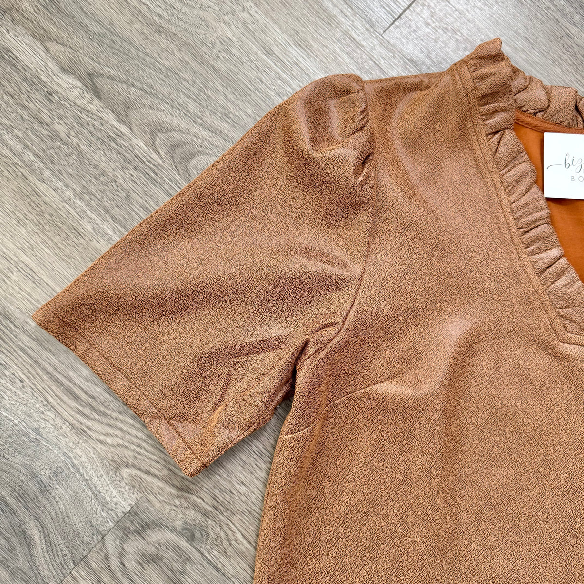 A close-up of a brown short-sleeve top with a ruffled V-neckline, laid flat on a gray wood-look floor. A white brand tag is visible inside the neckline.