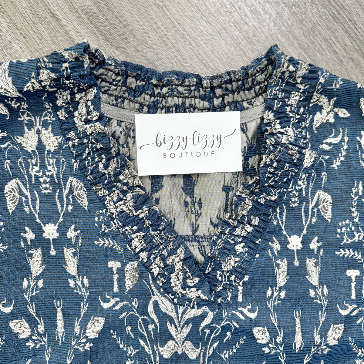 A close-up of a blue and white floral print blouse with a ruffled V-neckline, featuring a white brand label that reads 'bizzy bizzy BOUTIQUE'.