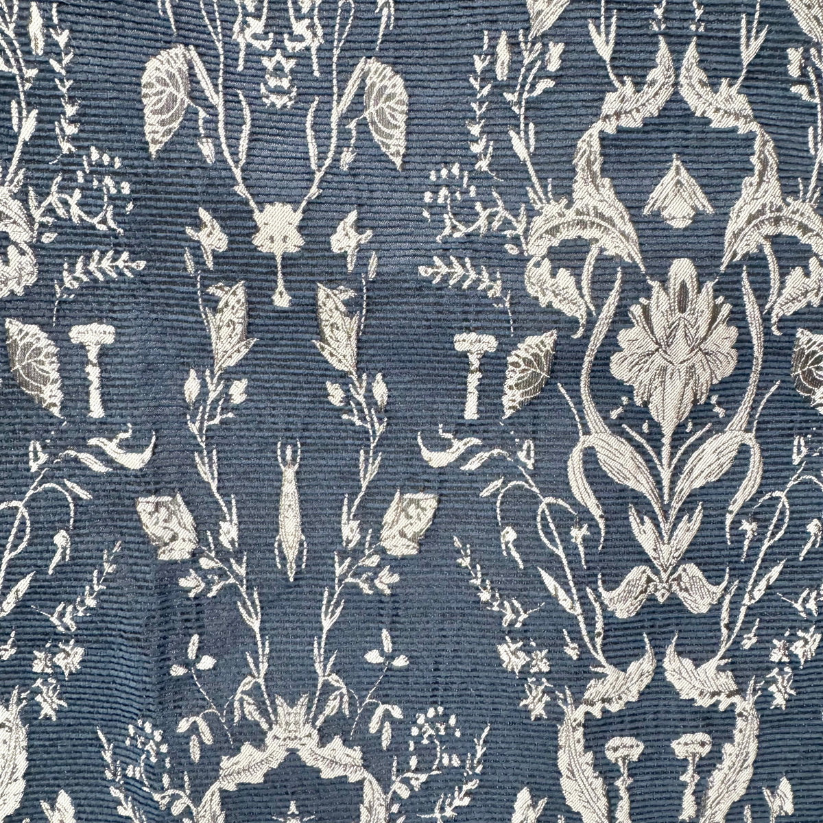 A close-up of a woven fabric pattern featuring a detailed botanical damask design in off-white on a textured slate blue background.