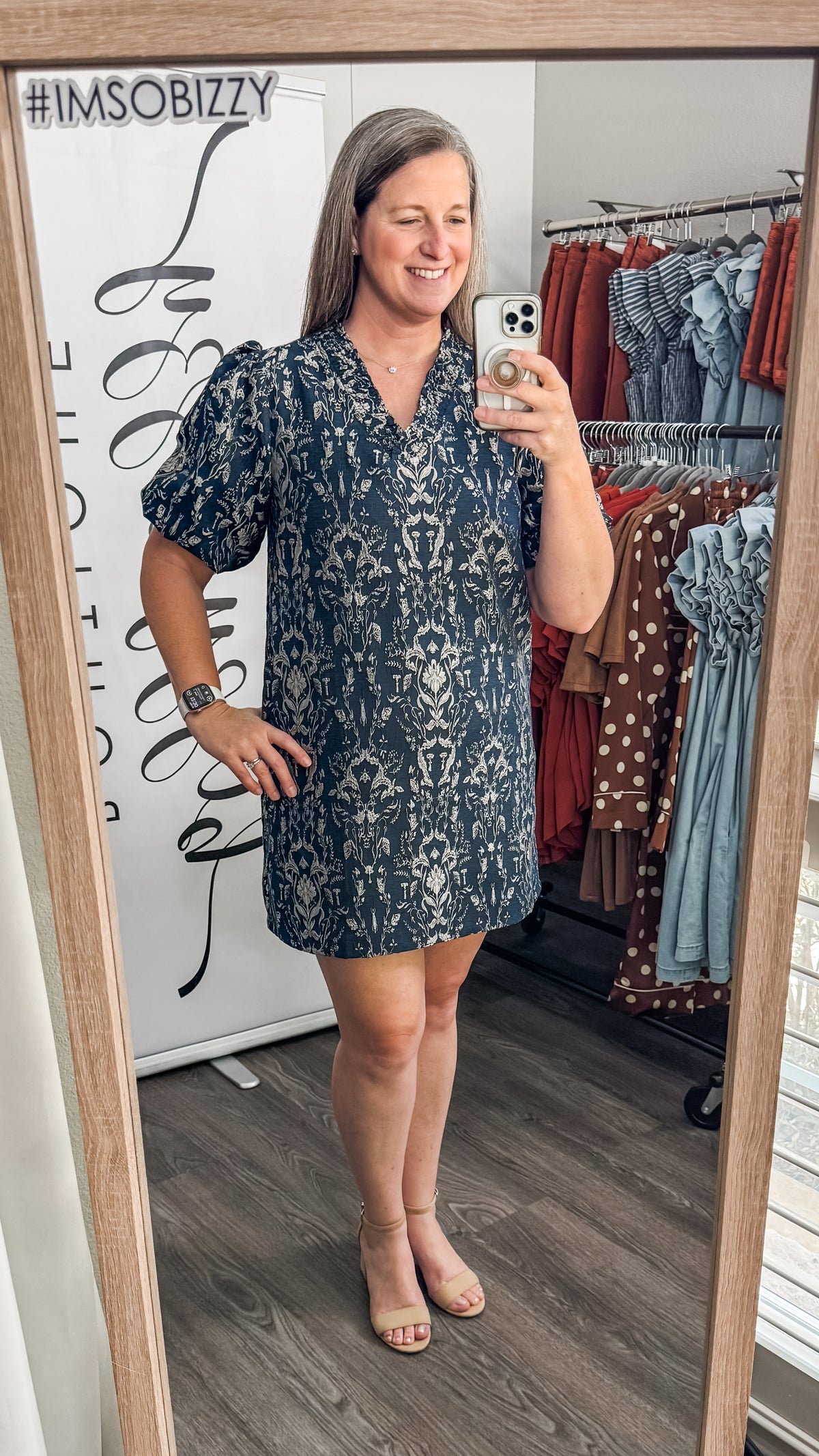 A woman takes a mirror selfie in a boutique wearing a short-sleeve navy blue and white patterned dress. The boutique interior is visible behind her, including a clothing rack with various garments and a white sign with black text.
