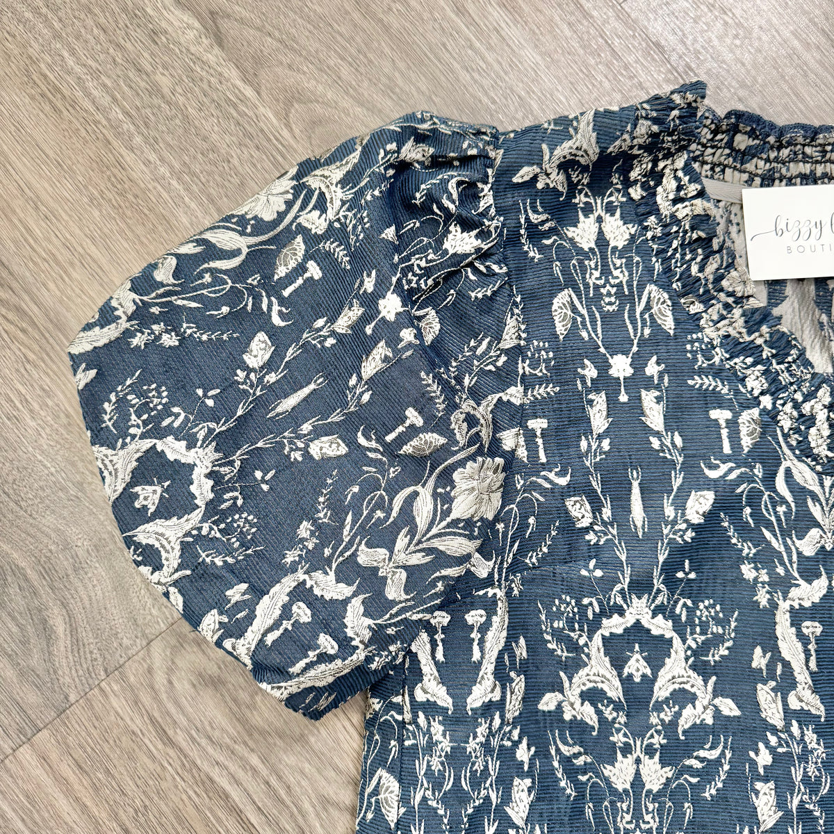 A close-up of a blue and white patterned blouse laid flat on a light wood floor. The blouse has a textured crinkle fabric, a ruffled smocked neckline, and short puff sleeves with frayed edges.