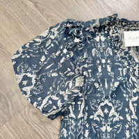 A close-up of a blue and white patterned blouse laid flat on a light wood floor. The blouse has a textured crinkle fabric, a ruffled smocked neckline, and short puff sleeves with frayed edges.