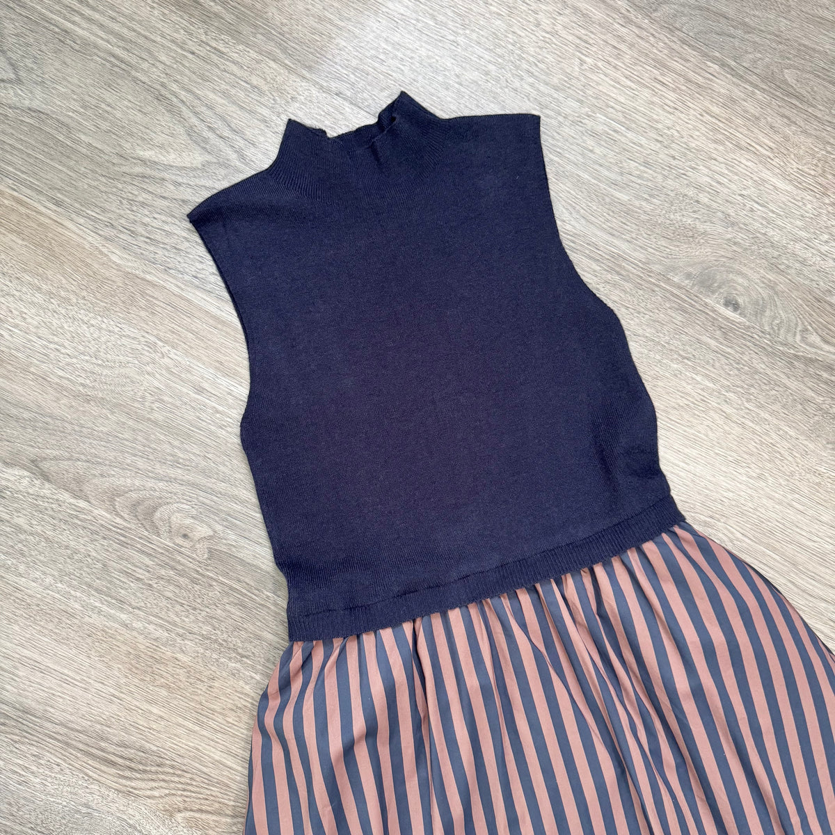 A sleeveless knit mock-neck dress in dark navy blue, paired with a skirt featuring vertical navy and brown stripes, laid flat on a light gray wood-look floor.