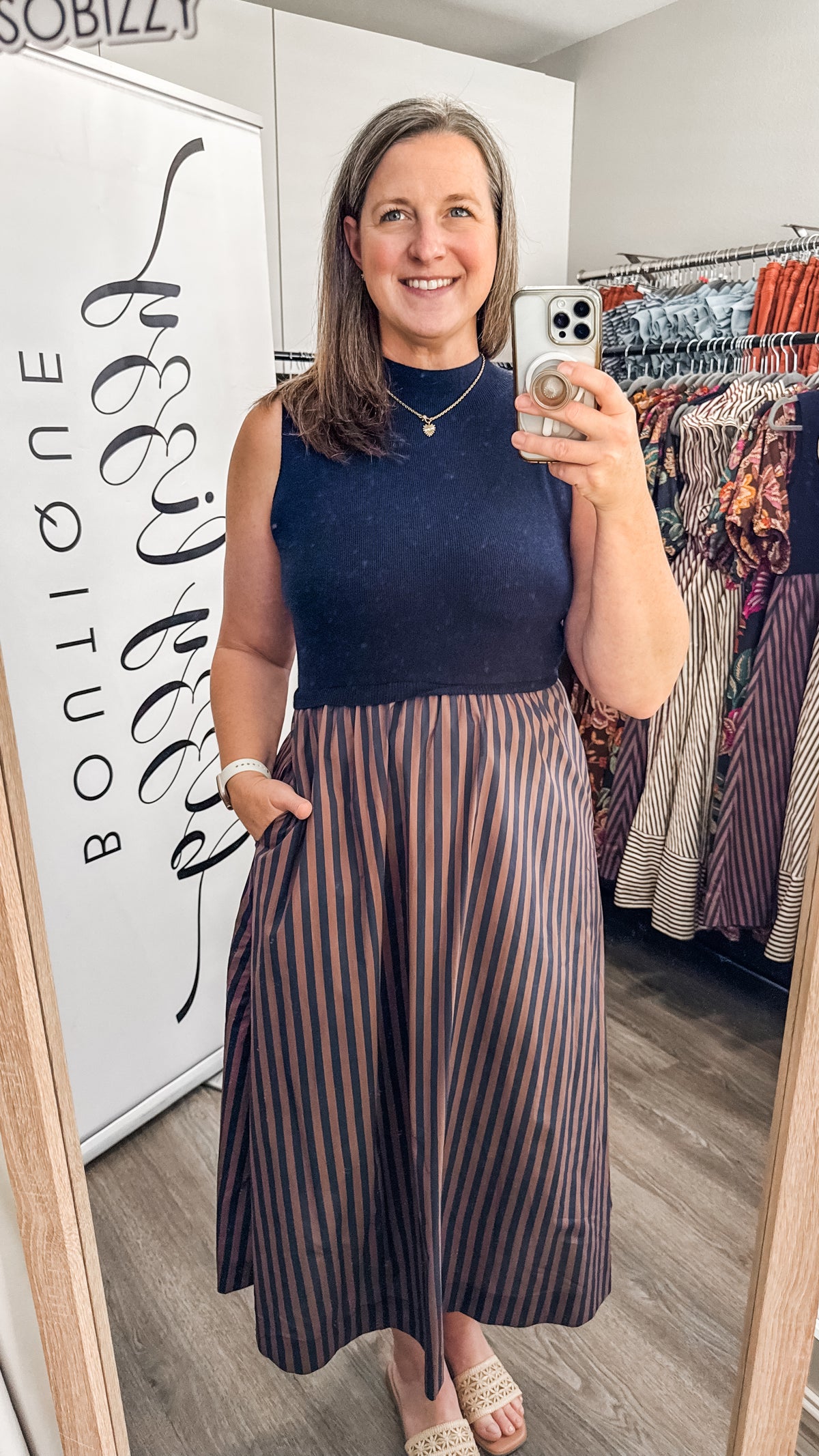 A woman takes a mirror selfie in a boutique wearing a sleeveless navy top and a pleated midi skirt with vertical stripes in navy, brown, and beige. A clothing rack with colorful garments is visible in the background.