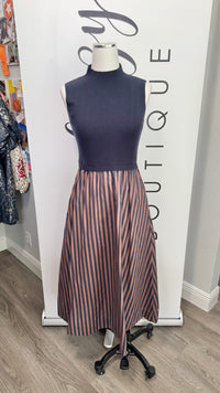 A sleeveless mock-neck dress displayed on a mannequin in a boutique setting. The top portion is solid navy blue, while the skirt features vertical stripes in navy, and brown.