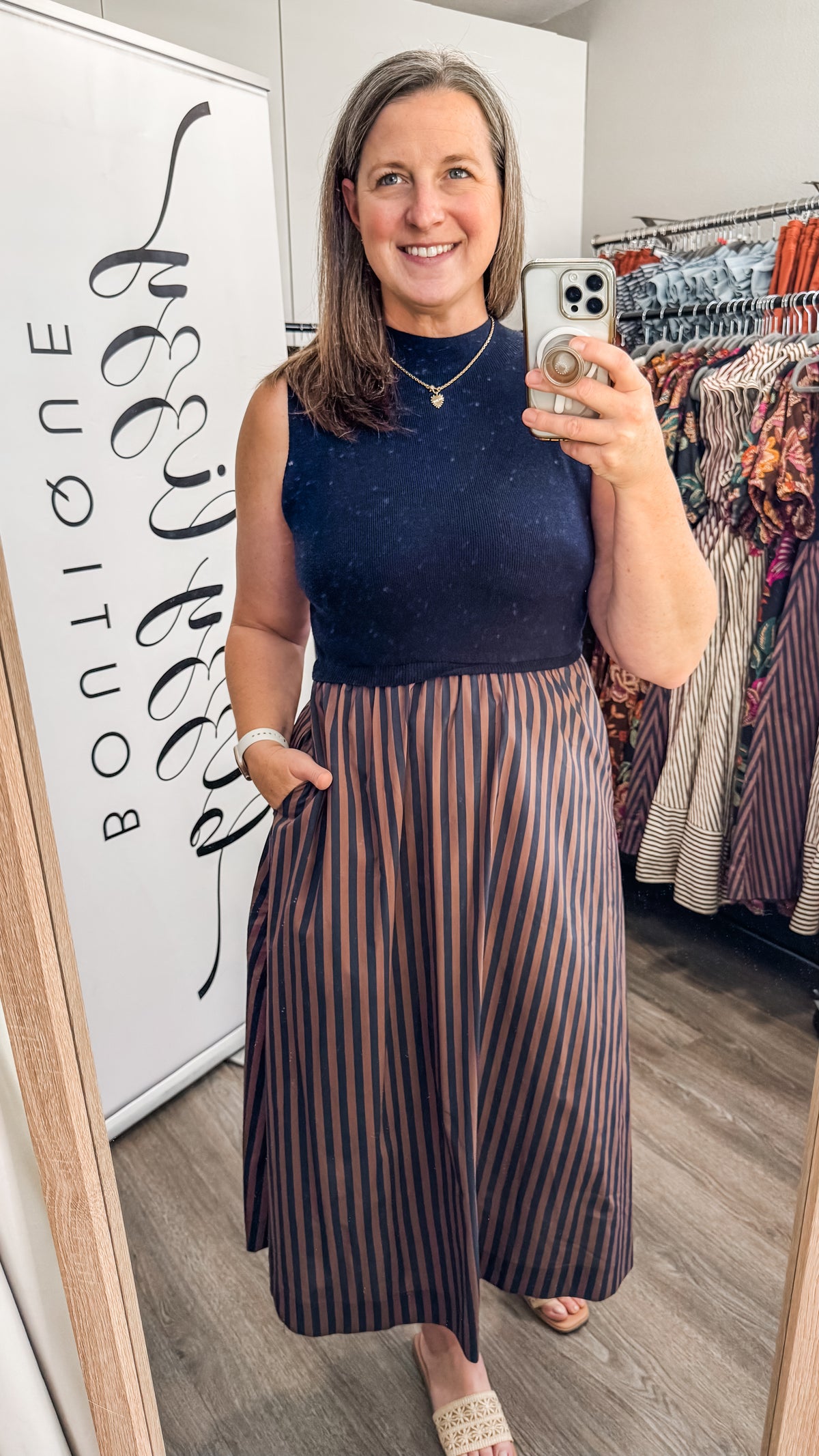 A woman takes a mirror selfie in a boutique wearing a sleeveless navy and brown dress with mock collar and striped navy and brown skirt. A white sign on the left reads 'BOUTIQUE'.