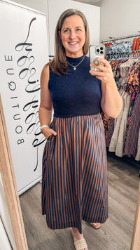 A woman takes a mirror selfie in a boutique wearing a sleeveless navy and brown dress with mock collar and striped navy and brown skirt. A white sign on the left reads 'BOUTIQUE'.