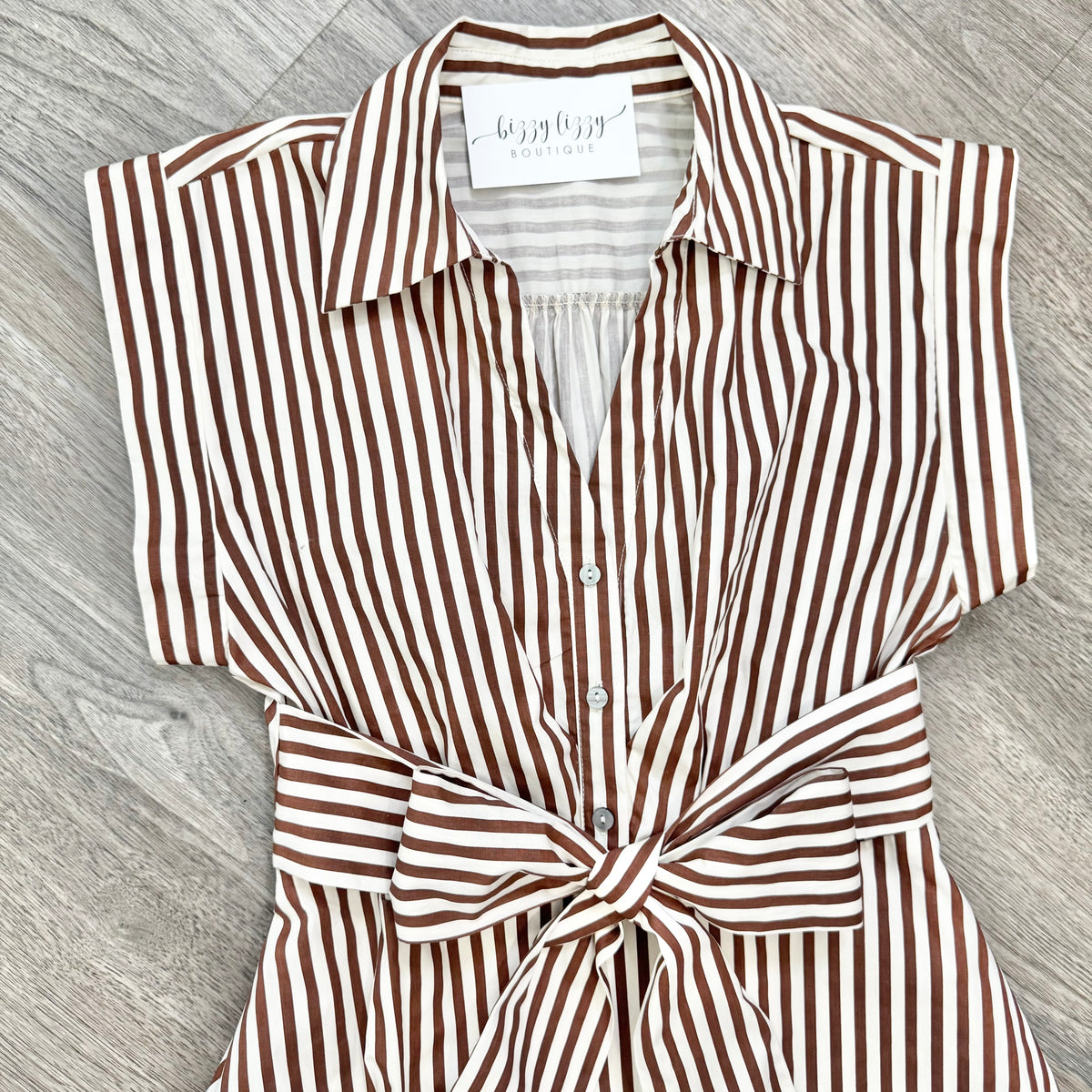 A brown and white vertically striped shirt dress with a tie waist, laid flat on a gray wood-textured surface. The brand name 'L'AGATHA' is visible on the inner neck label.