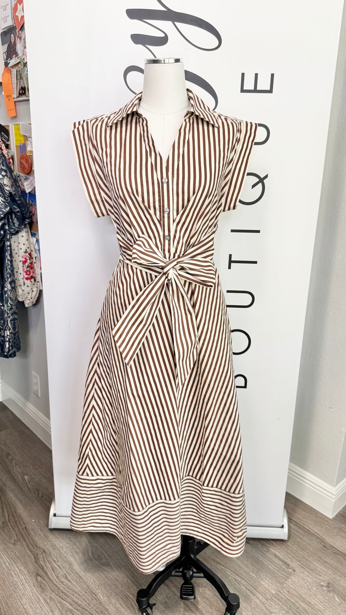 A short-sleeve collared dress with vertical brown and beige stripes, tied at the waist, displayed on a mannequin in front of a boutique banner.