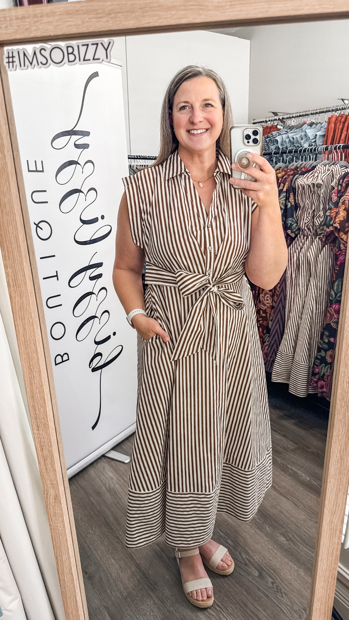 A woman takes a mirror selfie in a boutique while wearing a sleeveless, collared, belted midi dress with vertical brown and beige stripes. A white sign on the left reads '#IMSOBIZZY' and 'BOUTIQUE', and dress racks are visible in the background.