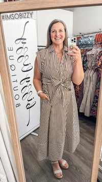 A woman takes a mirror selfie in a boutique while wearing a sleeveless, collared, belted midi dress with vertical brown and beige stripes. A white sign on the left reads '#IMSOBIZZY' and 'BOUTIQUE', and dress racks are visible in the background.