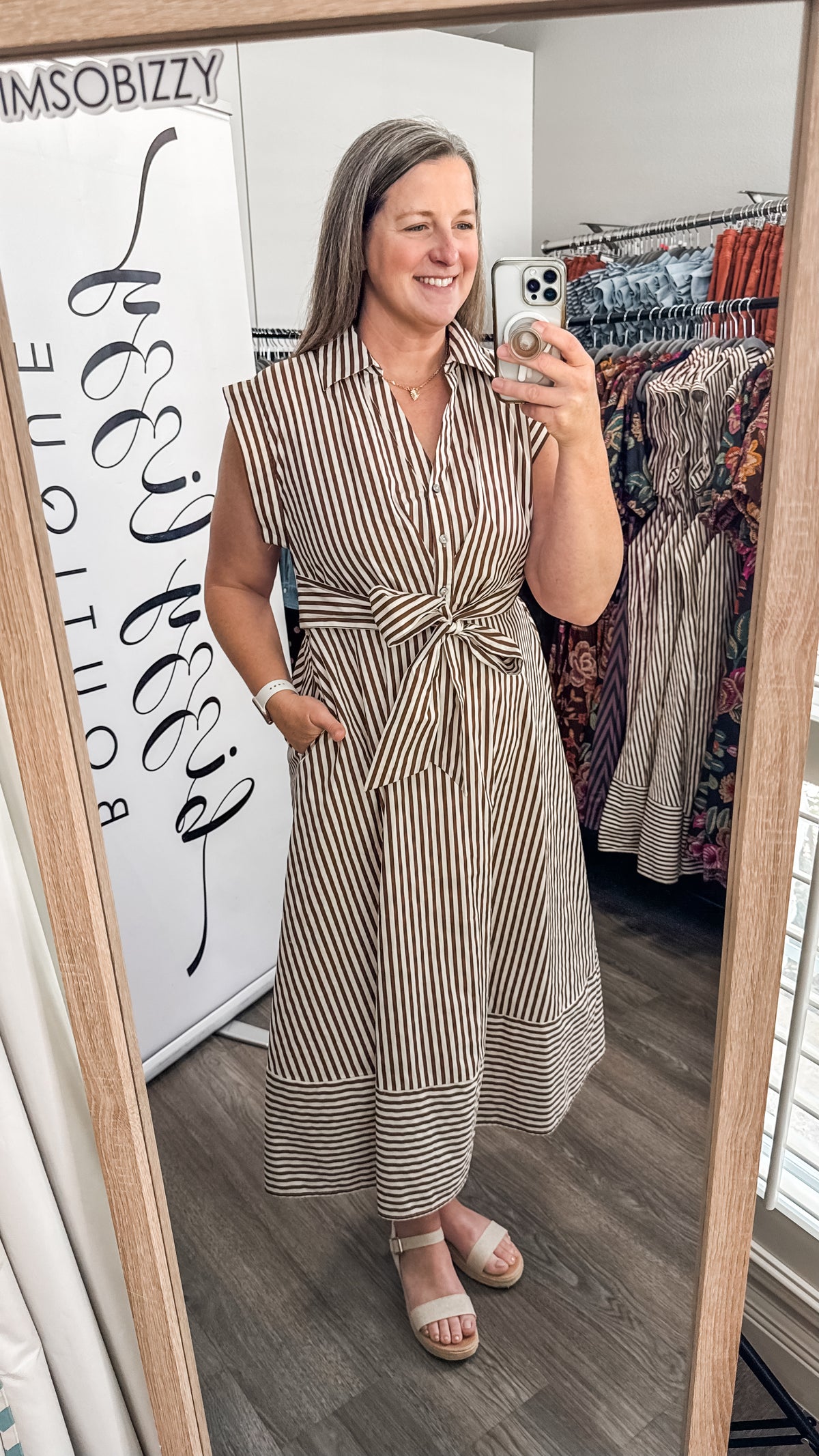 A woman takes a mirror selfie in a boutique wearing a sleeveless, button-front midi dress with vertical brown and white stripes and a tied waist. The boutique interior is visible, including a clothing rack with patterned dresses and a white sign reading 'IMSobizzy'.
