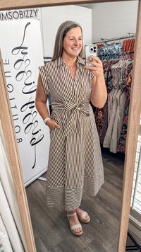 A woman takes a mirror selfie in a boutique wearing a sleeveless, button-front midi dress with vertical brown and white stripes and a tied waist. The boutique interior is visible, including a clothing rack with patterned dresses and a white sign reading 'IMSobizzy'.