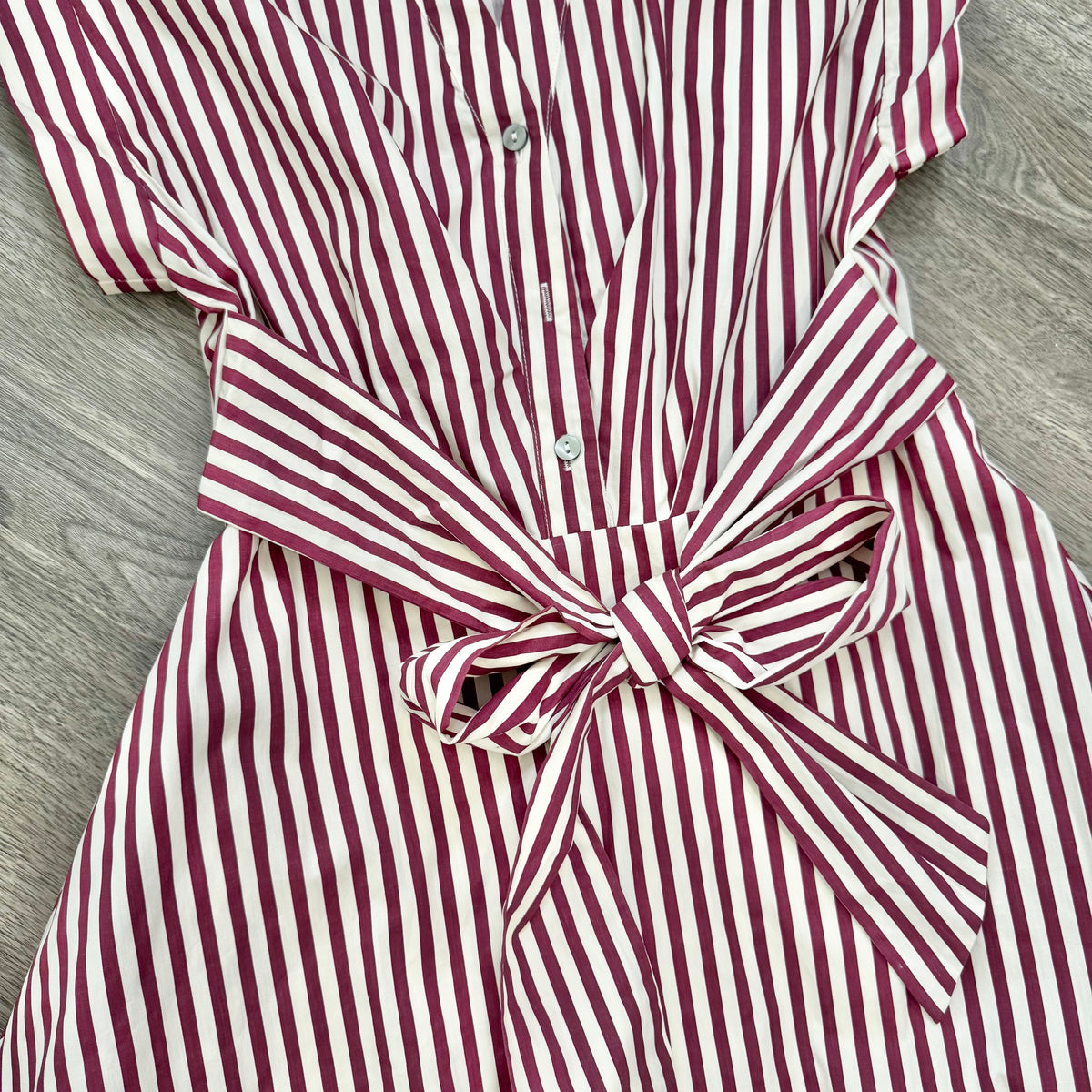 A close-up of a red and white vertically striped shirt dress with a matching fabric tie belt, laid flat on a gray wood-textured surface.