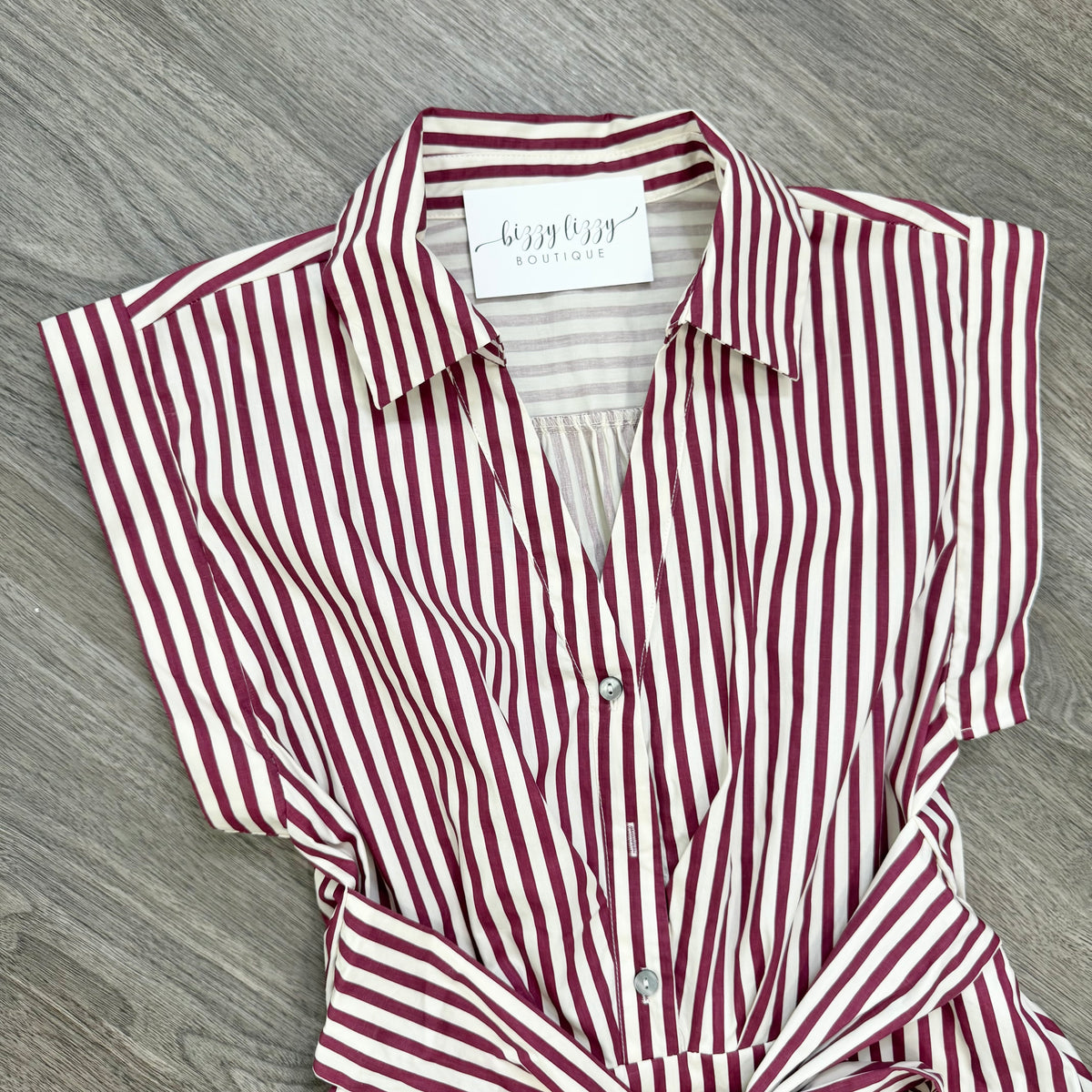 A burgundy and cream striped short-sleeve button-up shirt with a tie waist, laid flat on a gray wood-textured surface. The brand label reads 'Bizzy Lizzy Boutique'.