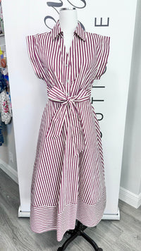 A sleeveless collared shirt dress with vertical burgundy and white stripes, featuring a tie waist and front pockets, displayed on a mannequin in front of a white boutique backdrop.