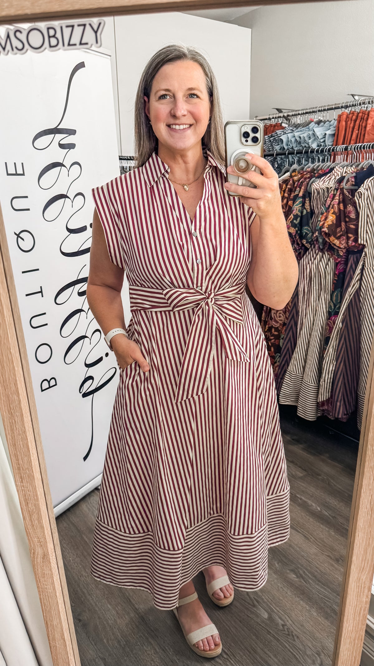 A woman takes a mirror selfie in a boutique wearing a sleeveless, button-front midi dress with vertical red and white stripes and a tied waist. Clothing racks and a boutique sign are visible in the background.