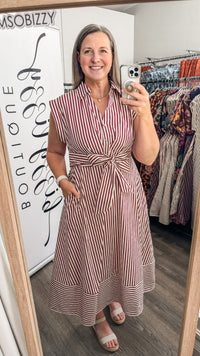 A woman takes a mirror selfie in a boutique wearing a sleeveless, button-front midi dress with vertical red and white stripes and a tied waist. Clothing racks and a boutique sign are visible in the background.