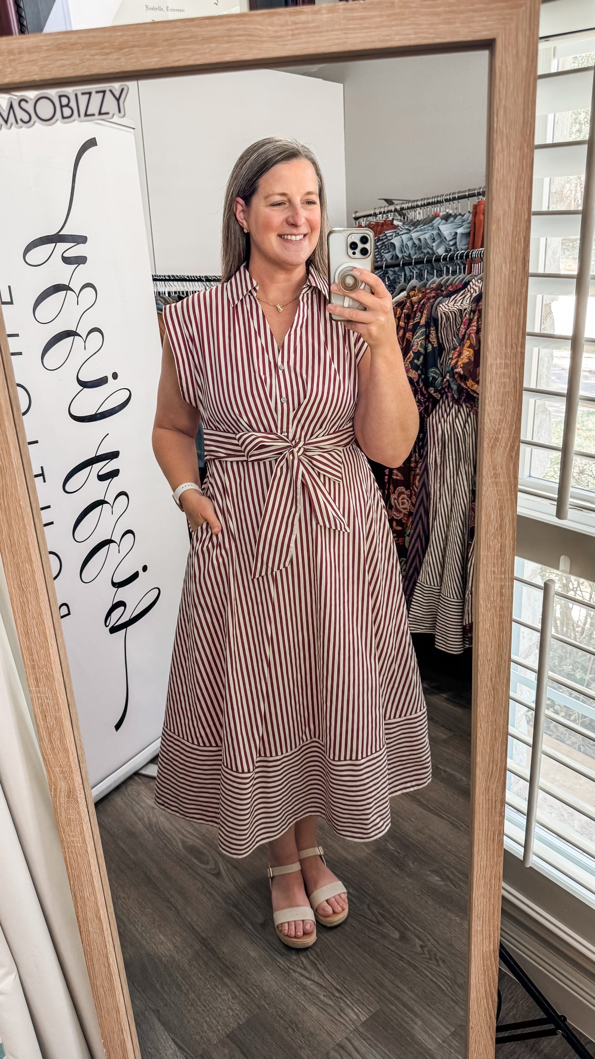 A woman takes a mirror selfie in a boutique wearing a sleeveless, button-front midi dress with vertical red and white stripes and a tied waist. The boutique interior is visible behind her, including clothing racks and a wooden-framed mirror.