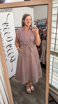 A woman takes a mirror selfie in a boutique wearing a sleeveless, button-front midi dress with vertical red and white stripes and a tied waist. The boutique interior is visible behind her, including clothing racks and a wooden-framed mirror.