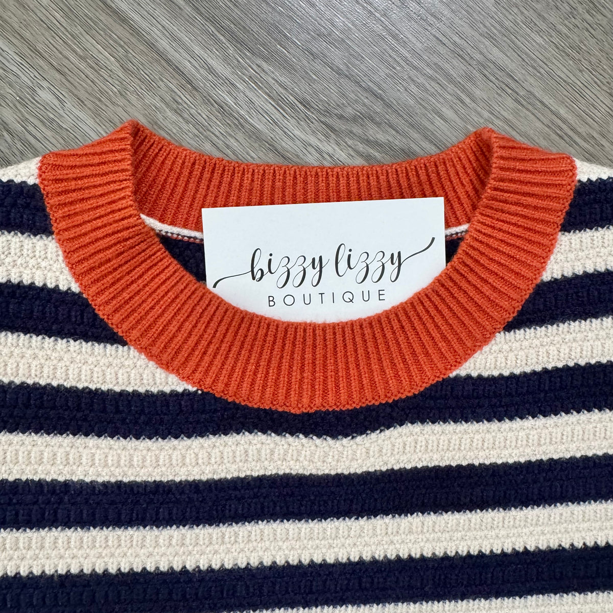 Close-up of a striped knit sweater with a rust orange crew neck, navy blue, and cream horizontal stripes, featuring a white brand label that reads 'bizzy lizzy BOUTIQUE'.