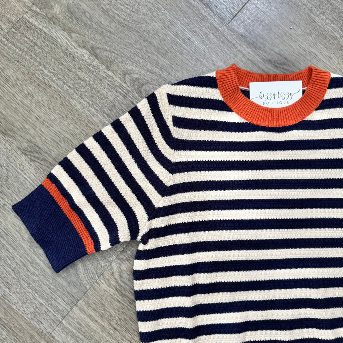 A knit sweater with horizontal navy and cream stripes, featuring a bright orange crew neck and sleeve cuff, laid flat on a gray wood floor. The brand label reads 'bizzy lizzy boutique'.