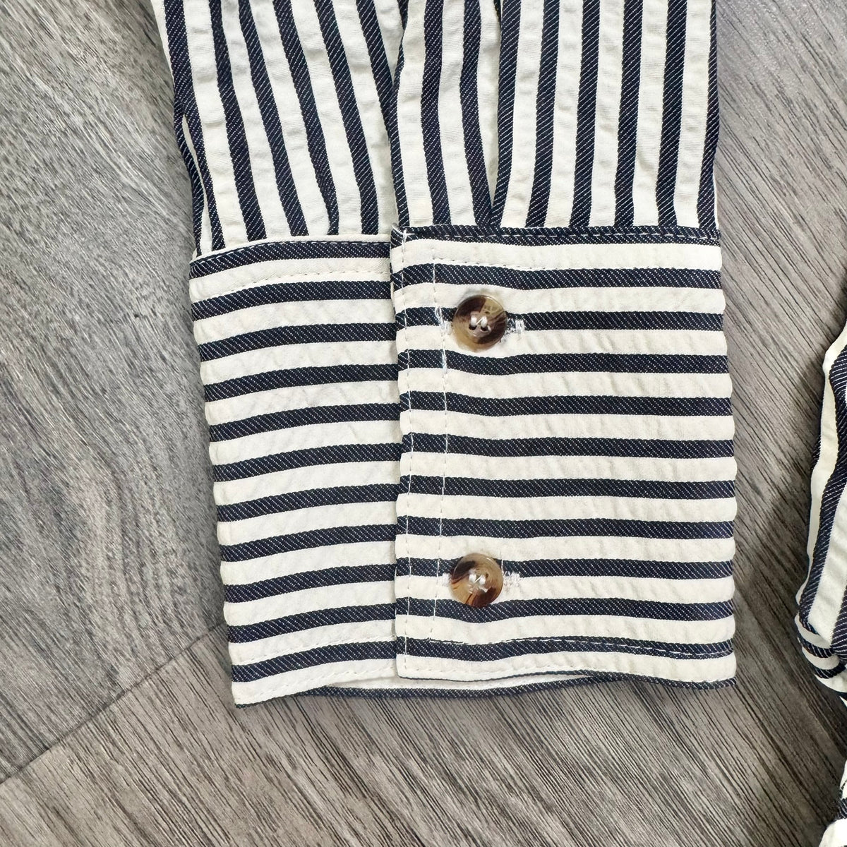 Close-up of a striped fabric garment, likely a shirt or blouse, showing navy and off-white horizontal stripes with two brown buttons on the cuff, laid flat on a gray wood-textured surface.