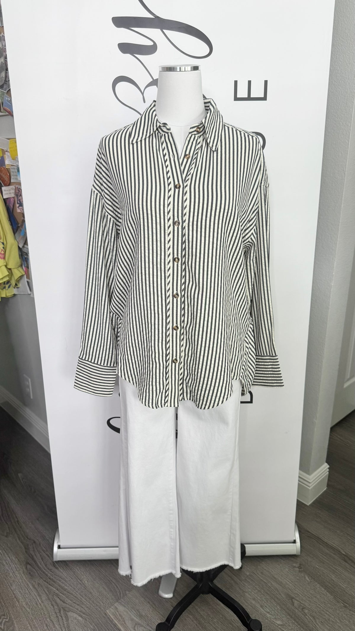 A long-sleeve button-up shirt with Dark blue and cream vertical stripes, styled on a white mannequin torso with white wide-leg pants against a white branded backdrop.