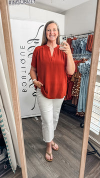 A woman takes a mirror selfie in a boutique fitting room, wearing a rust-orange short-sleeve V-neck top and white cropped pants. The boutique interior is visible behind her, including a clothing rack and a white sign with the brand name 'BONLIONE'.