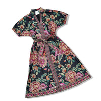 A short-sleeved wrap-style dress with a matching waist tie, featuring a vibrant floral print on a black background with pink, red, and green roses and leaves, finished with a decorative patterned border.