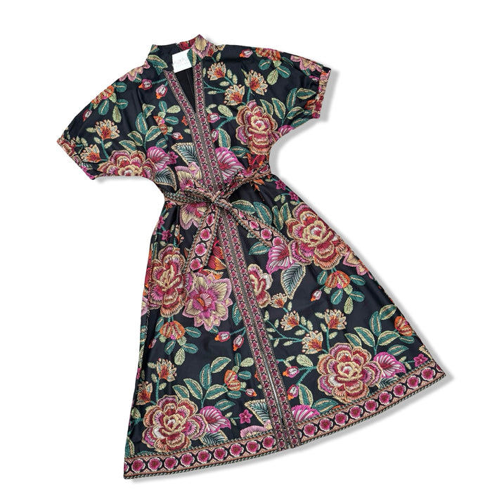 A short-sleeved wrap-style dress with a matching waist tie, featuring a vibrant floral print on a black background with pink, red, and green roses and leaves, finished with a decorative patterned border.