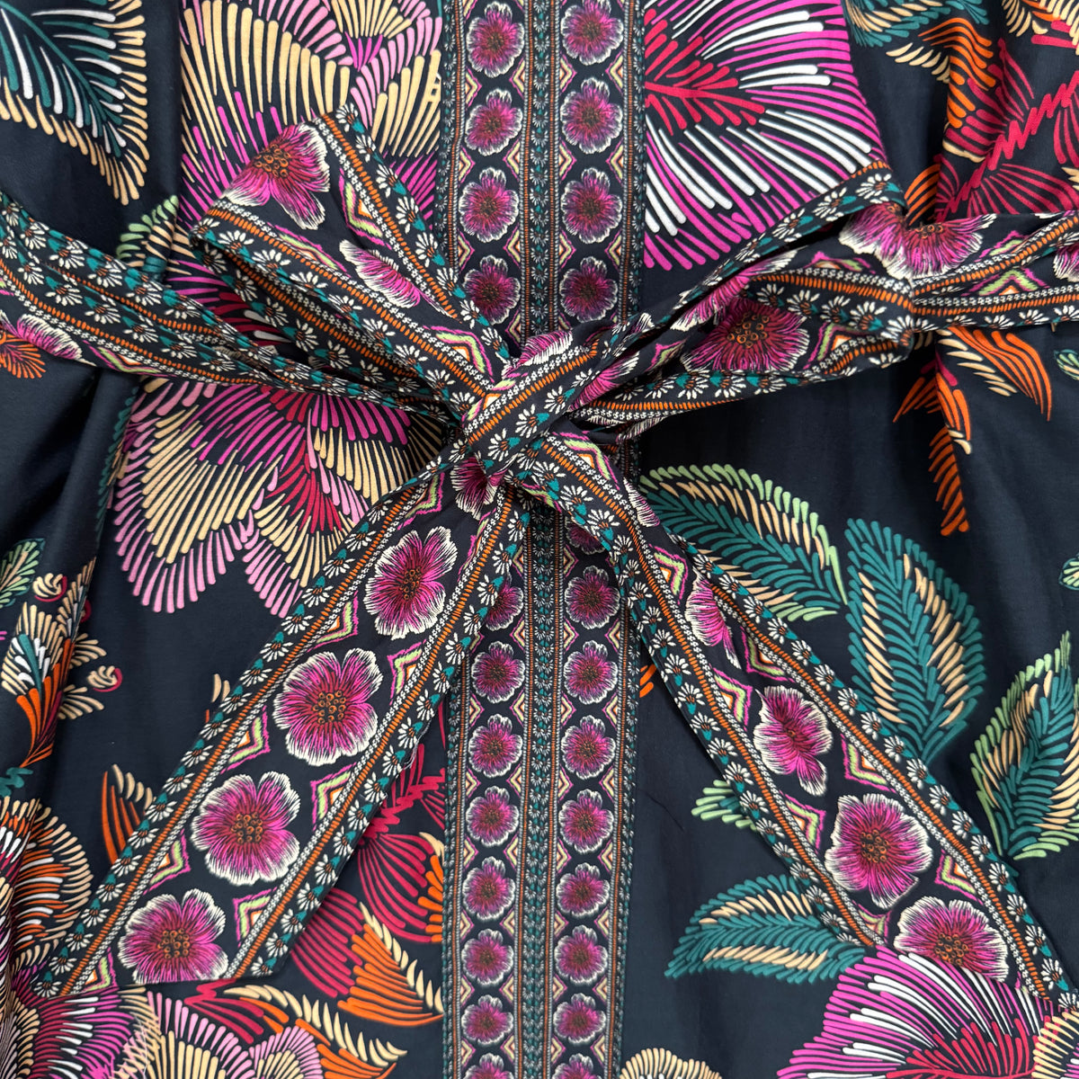 A close-up of a black patterned garment with a large tied waist sash. The fabric features a bold tropical floral and leaf print in pink, orange, teal, and cream, with a central vertical band of repeating pink floral motifs.