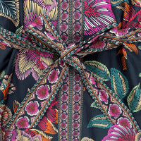 A close-up of a black patterned garment with a large tied waist sash. The fabric features a bold tropical floral and leaf print in pink, orange, teal, and cream, with a central vertical band of repeating pink floral motifs.