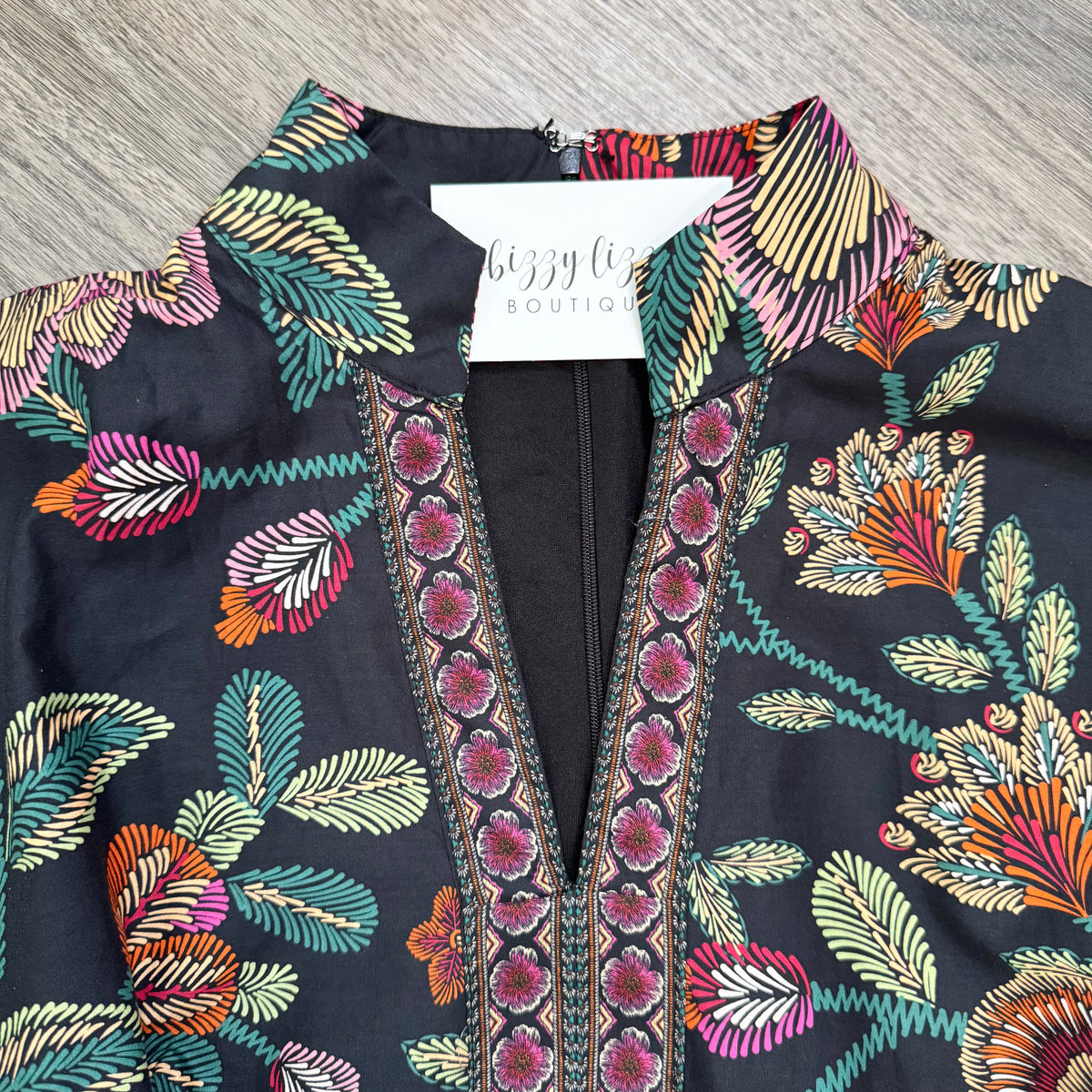 A close-up of a black embroidered blouse with a colorful floral and leaf pattern, featuring a split neckline with pink embroidered borders. A white brand tag reading 'bizzy life BOUTIQUE' is visible inside the collar, and the garment is laid flat on a gray wood-textured surface.