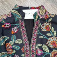 A close-up of a black embroidered blouse with a colorful floral and leaf pattern, featuring a split neckline with pink embroidered borders. A white brand tag reading 'bizzy life BOUTIQUE' is visible inside the collar, and the garment is laid flat on a gray wood-textured surface.