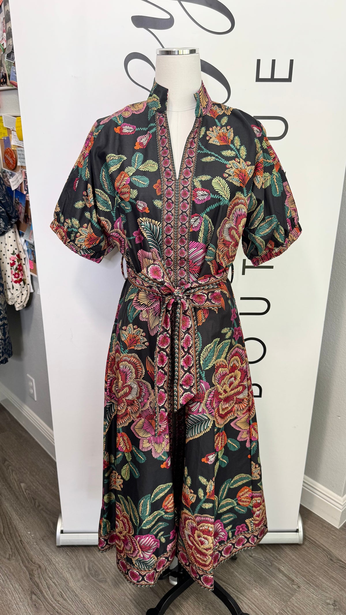 A short-sleeve wrap dress with a vibrant floral print on a black background, displayed on a mannequin in front of a white boutique backdrop.