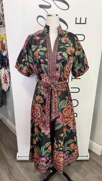 A short-sleeve wrap dress with a vibrant floral print on a black background, displayed on a mannequin in front of a white boutique backdrop.
