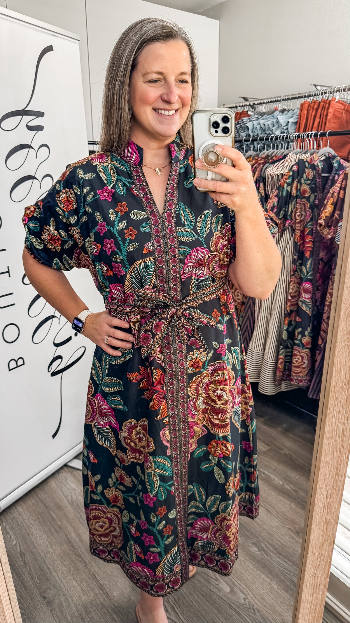 A woman takes a mirror selfie in a boutique wearing a dark green floral print dress with a tie waist and short sleeves. The dress features a vibrant mix of pink, red, orange, and yellow flowers with green leaves.