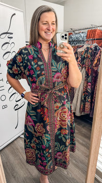 A woman takes a mirror selfie in a boutique wearing a dark green floral print dress with a tie waist and short sleeves. The dress features a vibrant mix of pink, red, orange, and yellow flowers with green leaves.