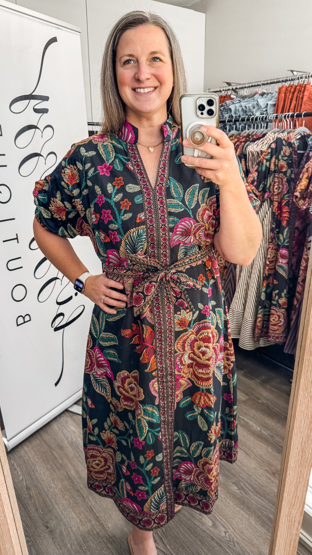 A woman takes a mirror selfie in a boutique while wearing a dark floral print tunic dress with a tie waist and puff sleeves. The dress features a black base with vibrant pink, red, and green floral patterns.