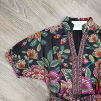 A short-sleeve kaftan-style top with a black base and a vibrant, multicolored floral and leaf pattern, laid flat on a gray wood-textured surface. The pattern features pink, orange, green, and yellow tones.