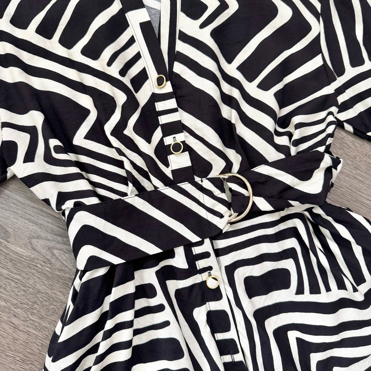 Black and white geometric patterned kimono-style top with a matching belt and gold-tone buttons, laid flat on a gray wood-textured surface.