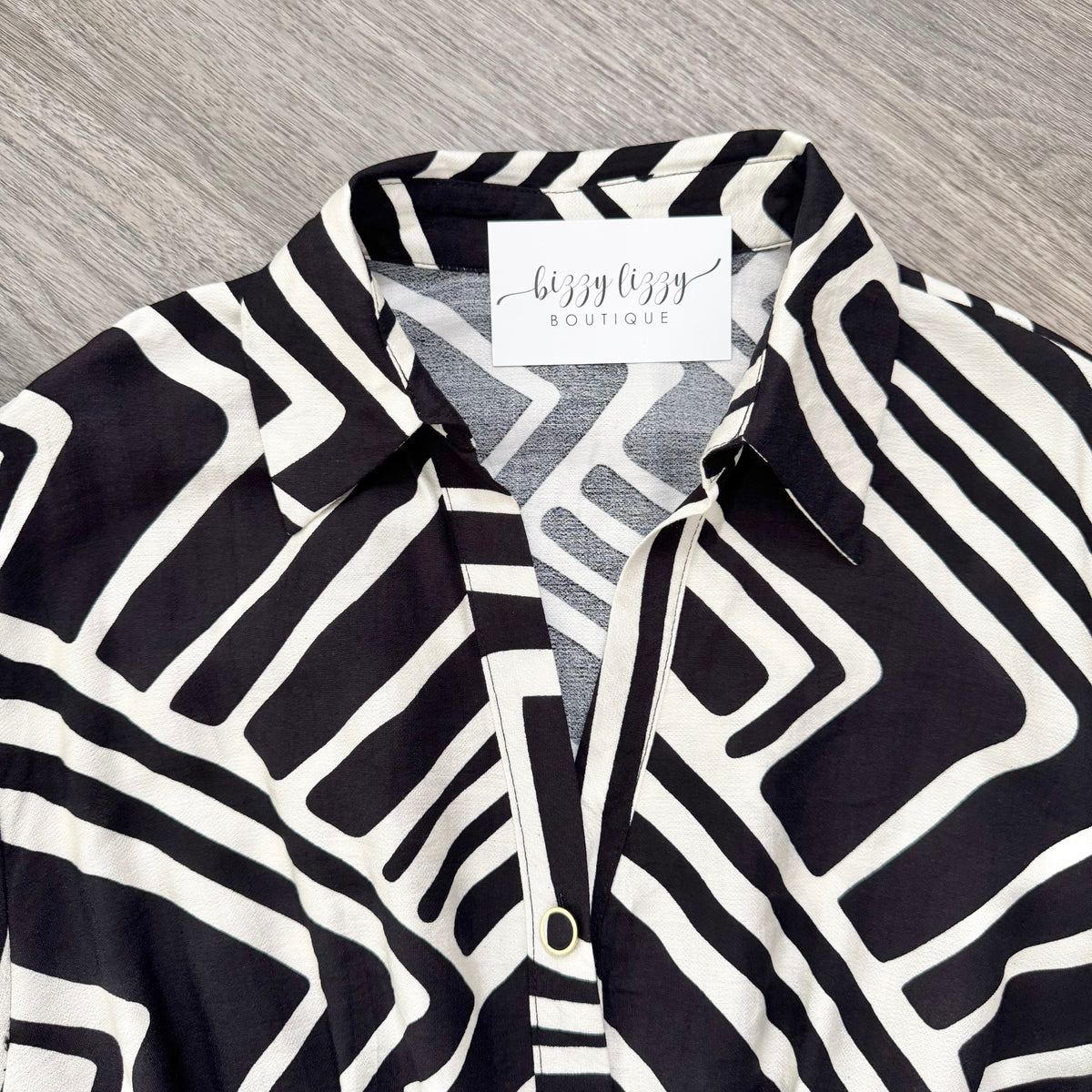 A close-up of a black and white geometric patterned button-down shirt with a visible 'bizzy bizzy boutique' neck label, laid flat on a gray wood-textured surface.