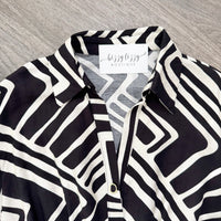 A close-up of a black and white geometric patterned button-down shirt with a visible 'bizzy bizzy boutique' neck label, laid flat on a gray wood-textured surface.