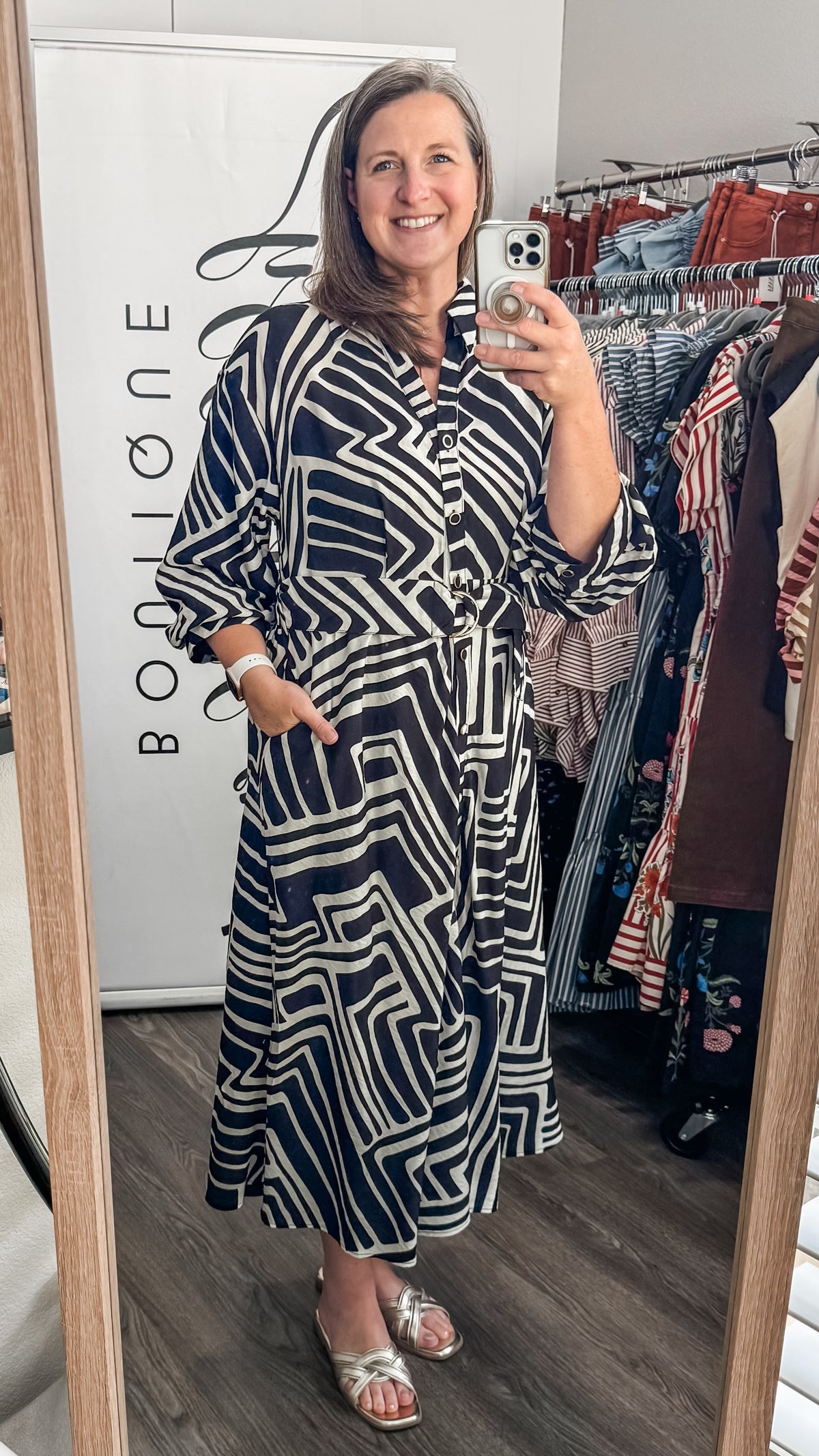 A woman takes a mirror selfie in a boutique showing a long-sleeve button-up dress with a bold black and cream geometric maze-like pattern. The boutique interior is visible, with a white wall with 'Bizzy Lizzy' text on the left and clothing racks on the right.