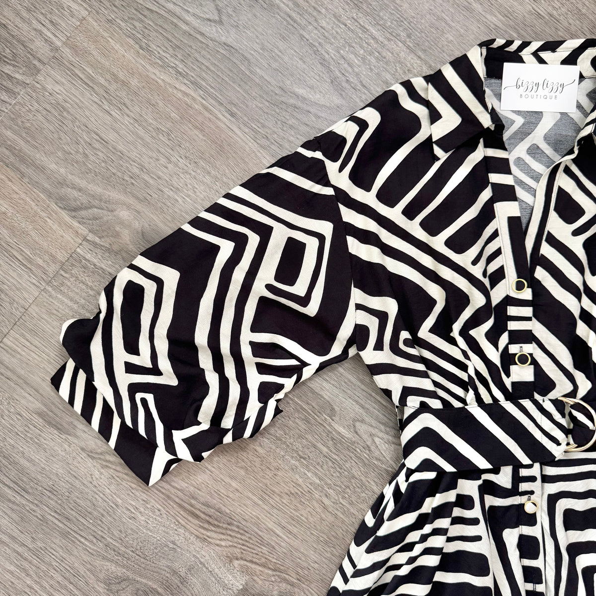 A black and white geometric patterned shirt dress with a collar, button front, and matching belt, laid flat on a gray wood-look floor. The brand label reads 'Bizzy Lizzy Boutique"
