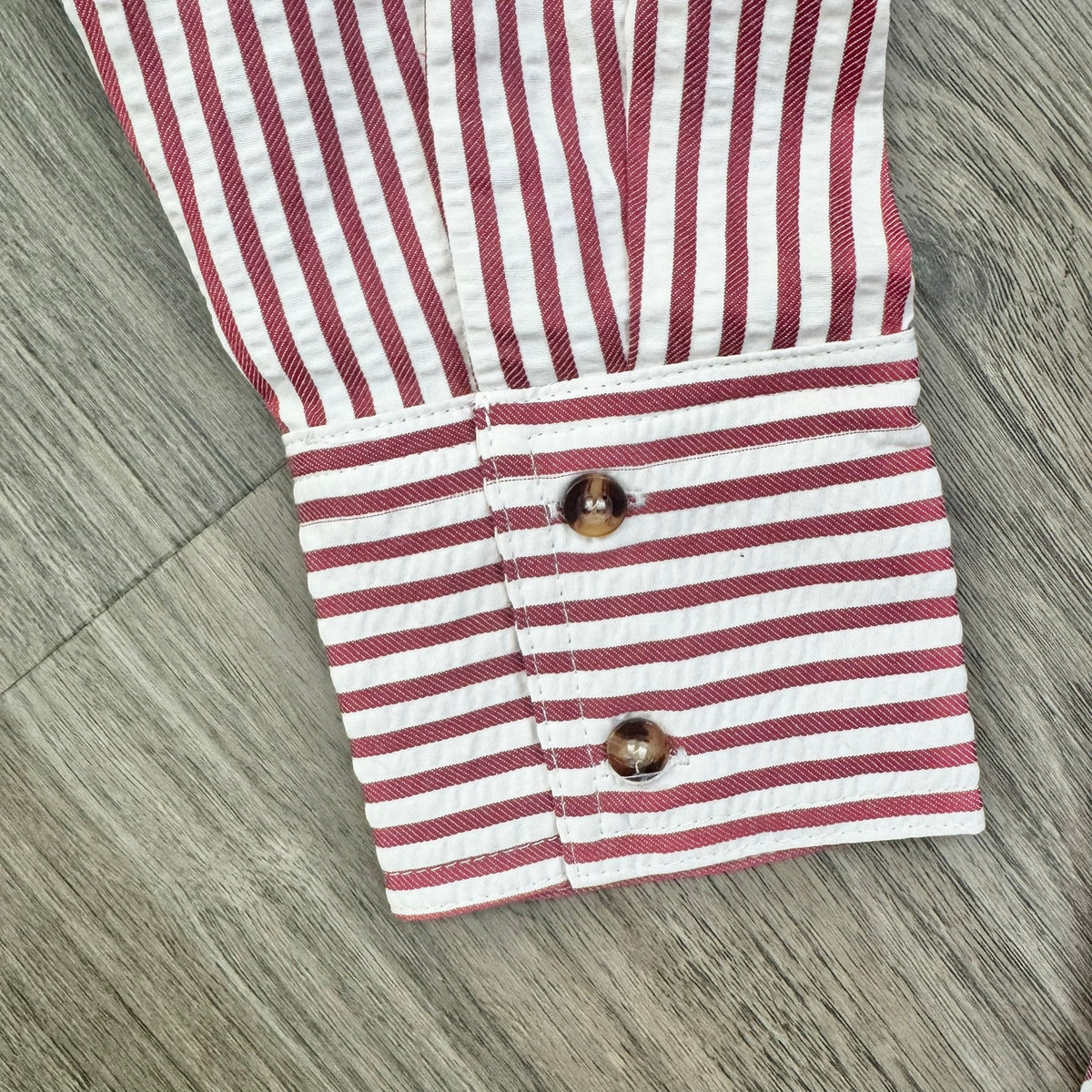 A close-up of a striped shirt cuff with red and white horizontal stripes and two brown buttons, laid flat on a gray wood-textured surface.